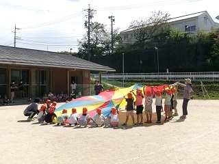 晴れた日に複数の子どもたちと数人の先生が丸いカラフルなシートの端を持っている様子