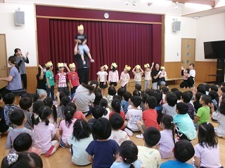 体育館で前列に頭に金の冠を乗せた子どもたちが並び、手前には座ってそれを観劇している数十人の子どもたちがいる様子