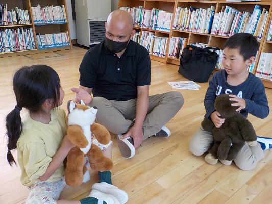 図書館のようなところで大人の左右に動物の人形を抱えた子どもたちが一人ずつ座り指導を受けている様子