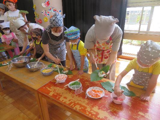 三角巾とエプロンを身に着けた子どもたちが大人と一緒に料理を作っている様子