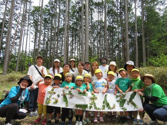 複数の親子と先生が揃ってポーズを取っている様子。前には「おやこえんそく」と草花で縁取られた大きな紙が写っている。