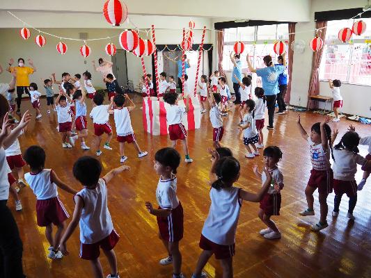 室内で複数の子どもたちが円状になって踊っている様子。中心には櫓がある。