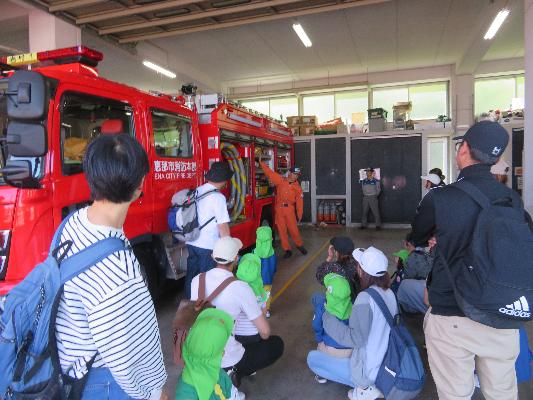 複数の子どもたちと大人が消防車のそばに座り消防署員から説明を受けている様子